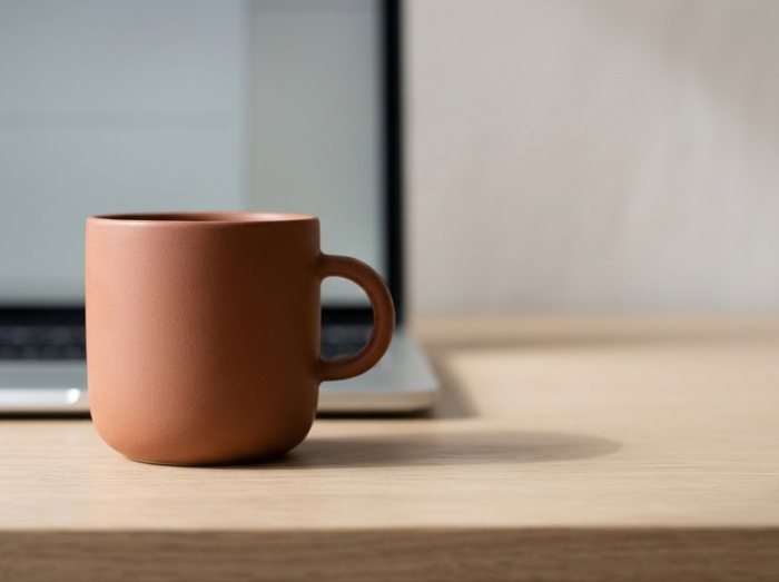 The terracotta ceramic mug in close focus on the pale oak desk, the laptop softly out of focus behind it.