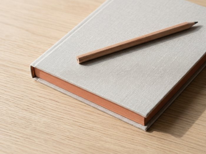 Macro detail of the linen-bound notebook and wood pencil on the oak desk, the terracotta-stained page edge catching the light.